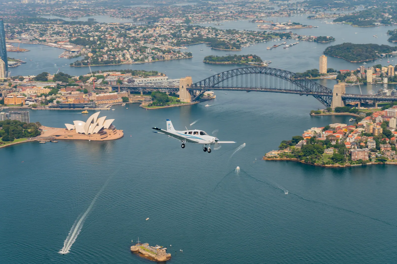 A training aircraft flying over Sydney Harbour, with the Opera House and the Harbour Bridge below